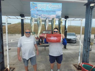 Two people wearing sunglasses stand under a fishing charter sign with five fish hanging from a rack behind them.