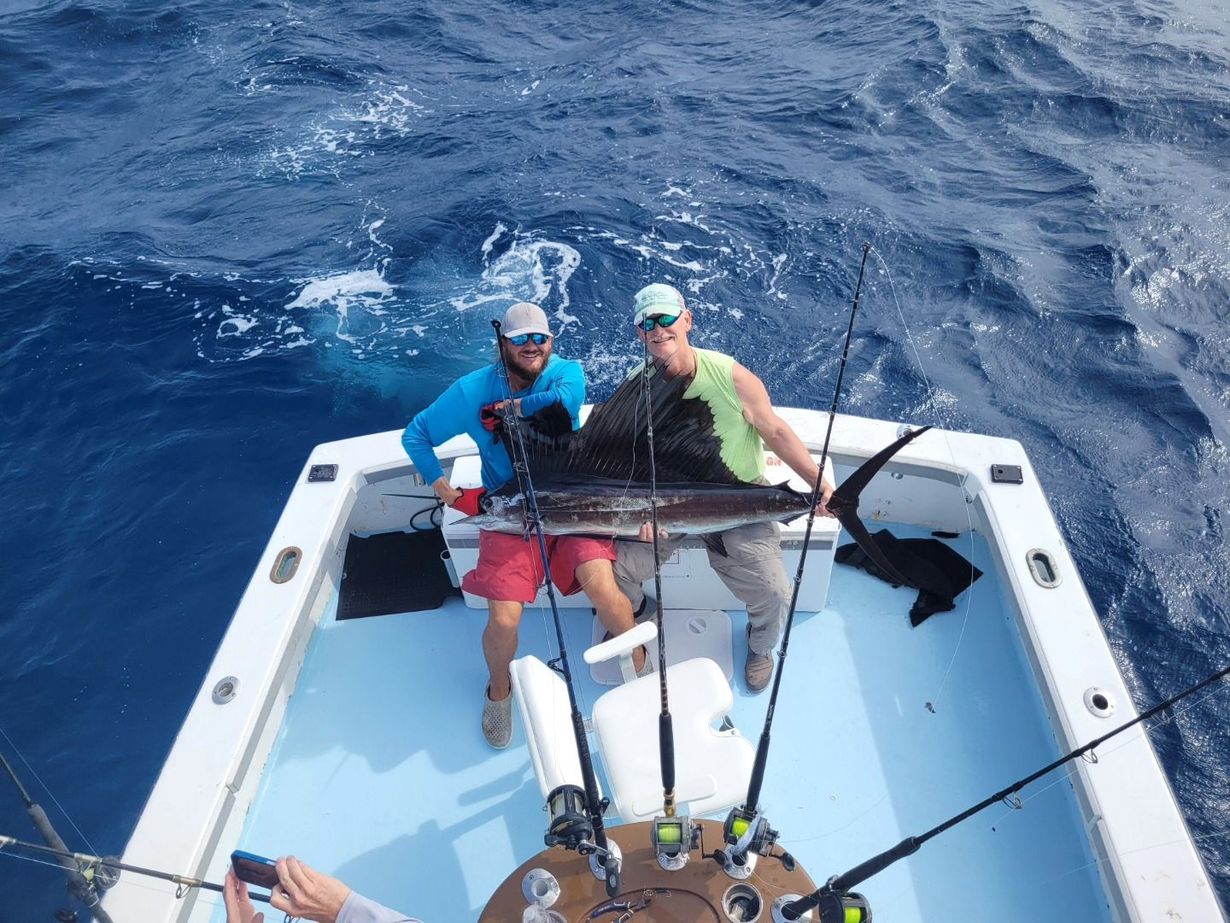 Two people sit on the deck of a boat holding a large sailfish caught while fishing in deep blue water.