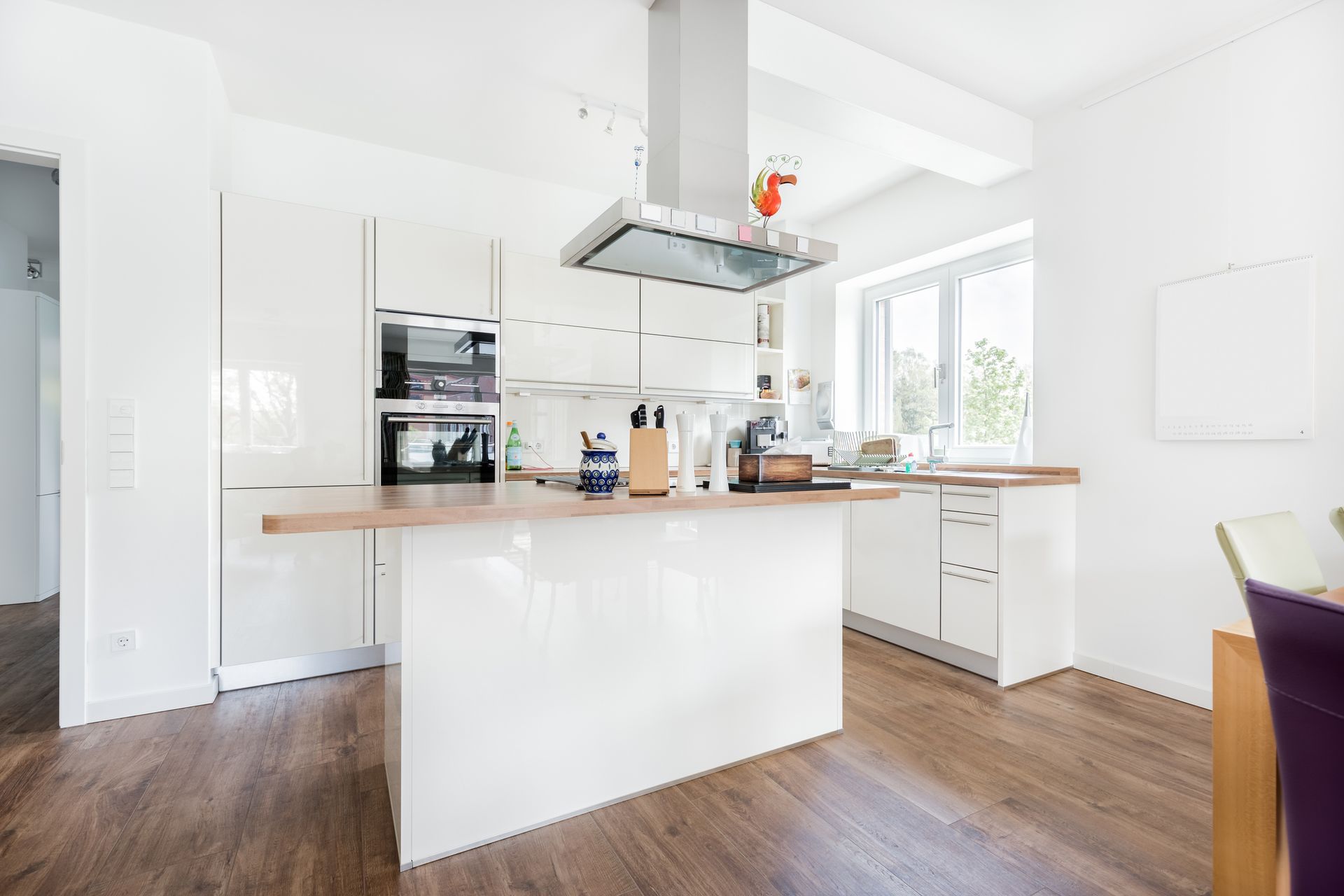 Modern white kitchen with wooden floors and countertops. It features an island, stainless steel range hood, and lots of natural light.