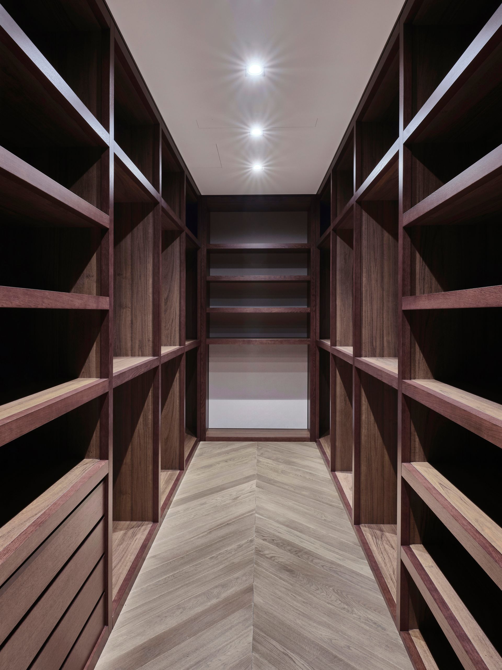 Dark wood walk-in closet with shelves on both sides, chevron-patterned floor, and recessed lighting. Empty and well-lit.