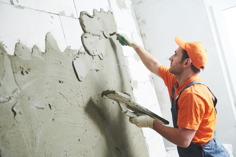 Man Wearing an Orange Shirt and Cap Plastering a Wall With a Trowel, Wearing Gloves — Rowal Constructions in Kotara, NSW