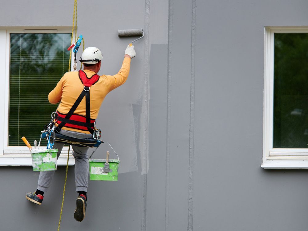 Painter Using a Roller on a Wall While Suspended by Ropes and Safety Equipment — Rowal Constructions in Cardiff, NSW