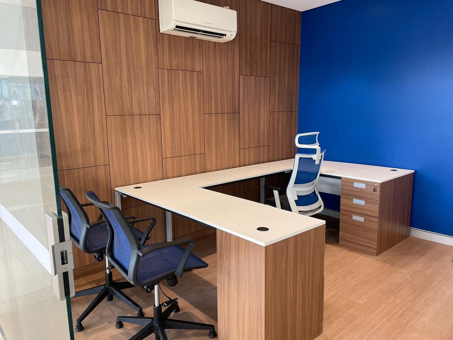 L-shaped office desk with two blue chairs. Wooden panel wall and blue wall. An air conditioner is above the desk.