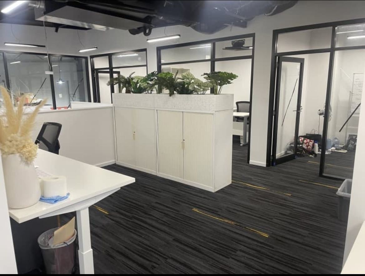 Office interior with white desks, cabinets, and glass-walled rooms. Plants decorate the space.