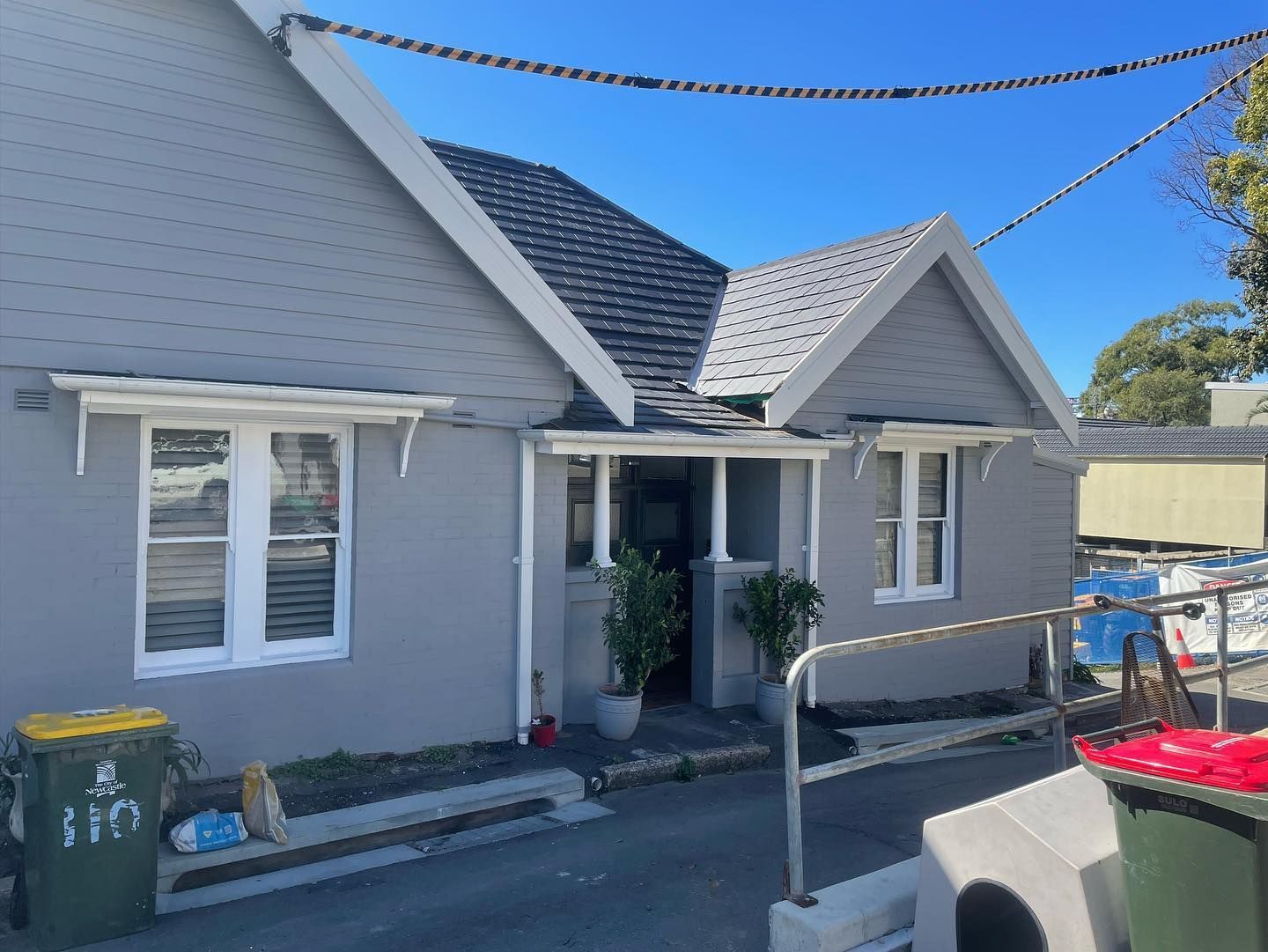 Gray house with white trim, dark roof, front porch, two windows, trash cans, and a ramp.