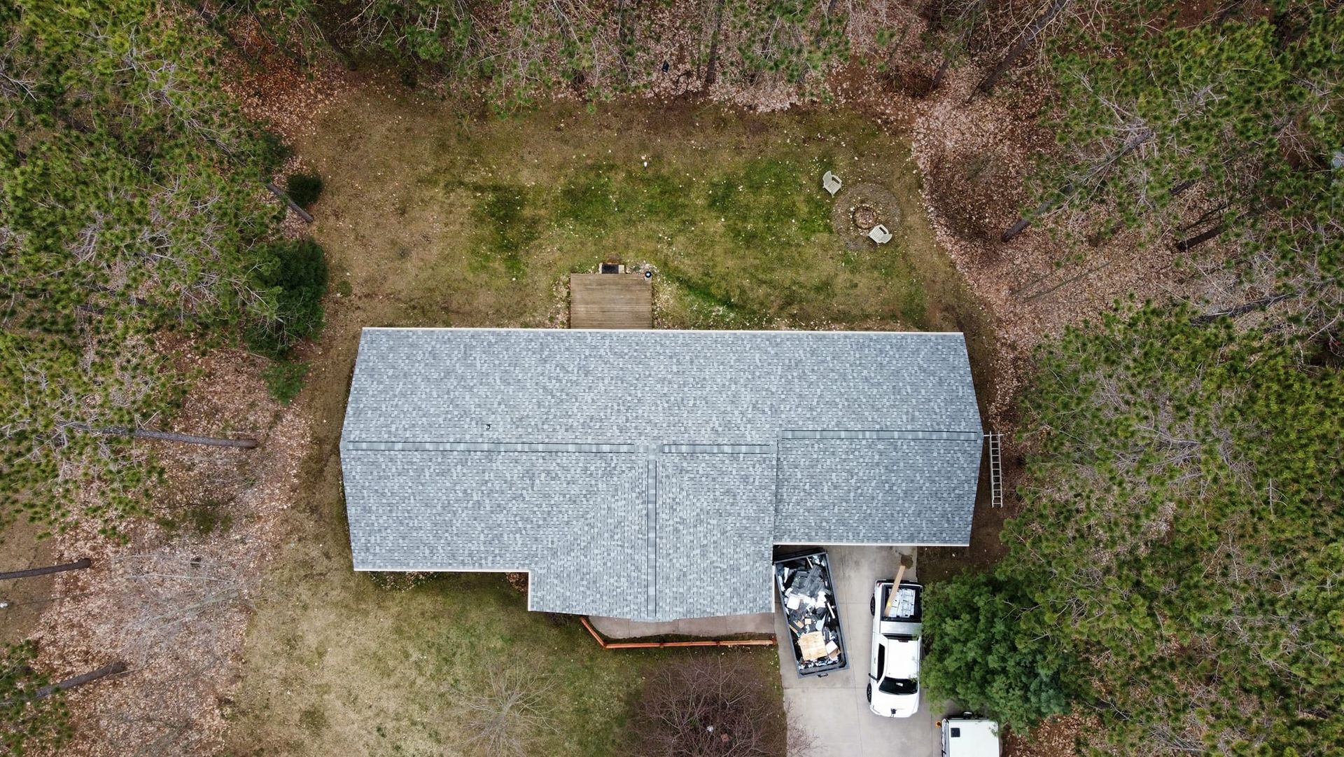 Aerial view of a house with a gray shingled roof, surrounded by trees, with two parked cars in the driveway.