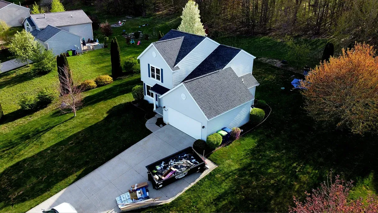 Aerial view of a white suburban house with a large dumpster on the concrete driveway and green lawn.