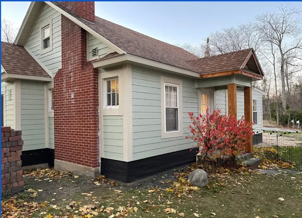 A light green, one-story house with a brick chimney and a small, wood-pillared porch under a brown shingled roof.