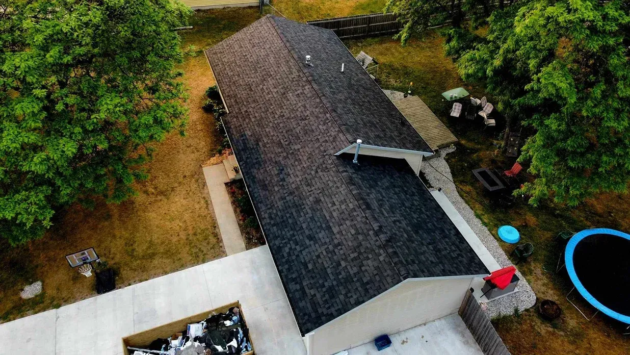 Aerial view of a house with a dark shingled roof, surrounding trees, a concrete patio, and a backyard trampoline.