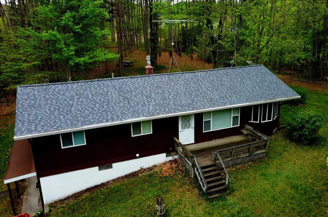 An aerial view of a single-story house with dark siding, a gray shingled roof, and a wooden deck, set in a wooded area.