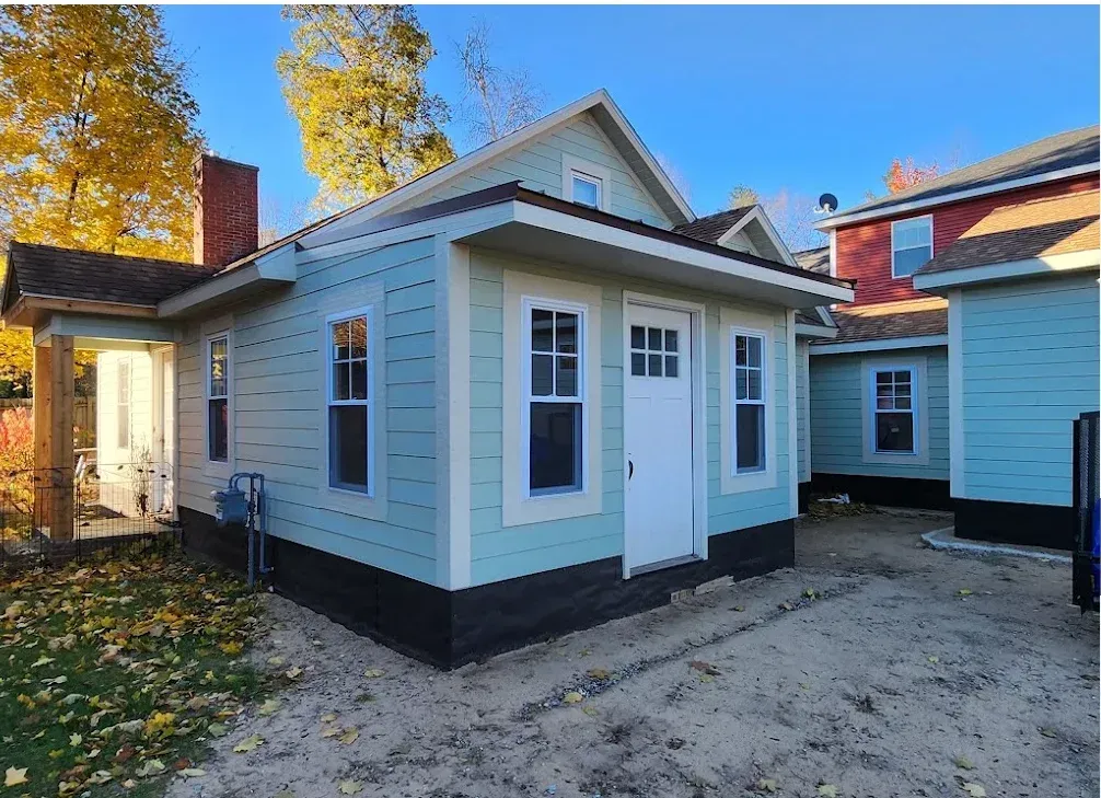 A light blue, newly constructed small house with a white door and dark foundation, surrounded by autumn leaves.
