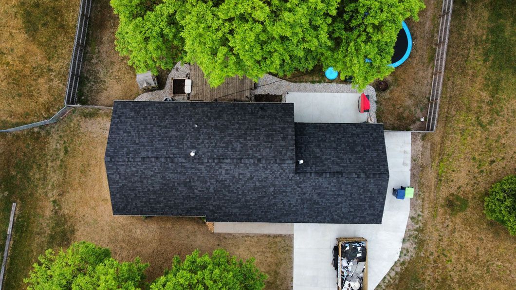 A top-down aerial view of a dark-roofed house with concrete walkways, surrounded by trees and a fenced yard.