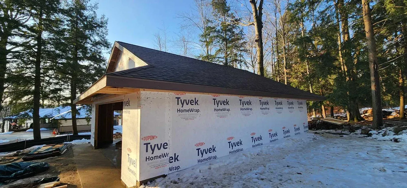 A small, unfinished building with a brown roof and white protective house wrap, set in a snowy, wooded outdoor area.