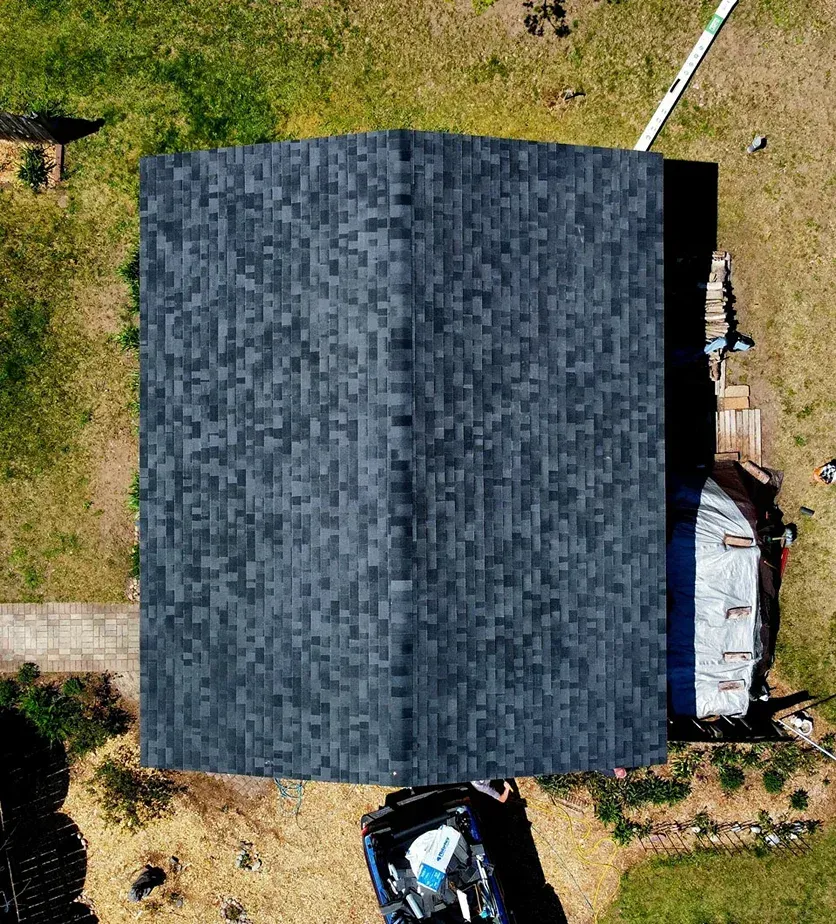 Aerial view of a rectangular house roof with dark gray shingles, surrounded by a grassy lawn and a parked vehicle.