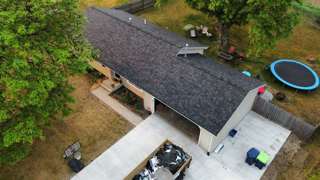 Aerial view of a residential home with a dark shingled roof, a concrete driveway, and a backyard with a trampoline.