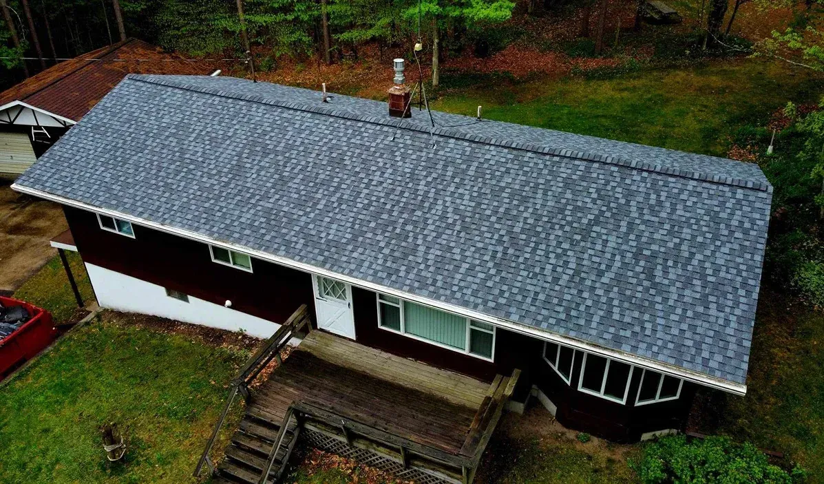 An aerial view of a single-story house with a new grey shingled roof, dark brown siding, and a wooden front deck.