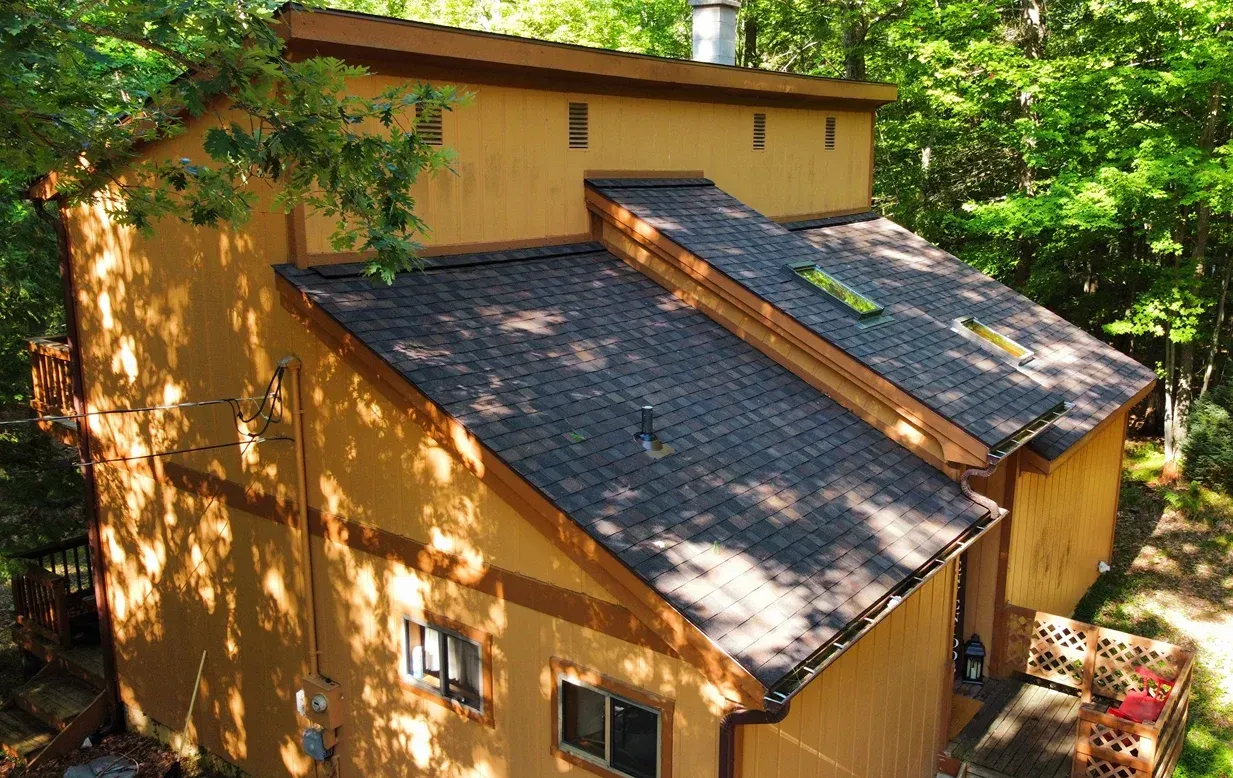 A multi-level tan house with a dark shingled roof nestled among dense green trees.