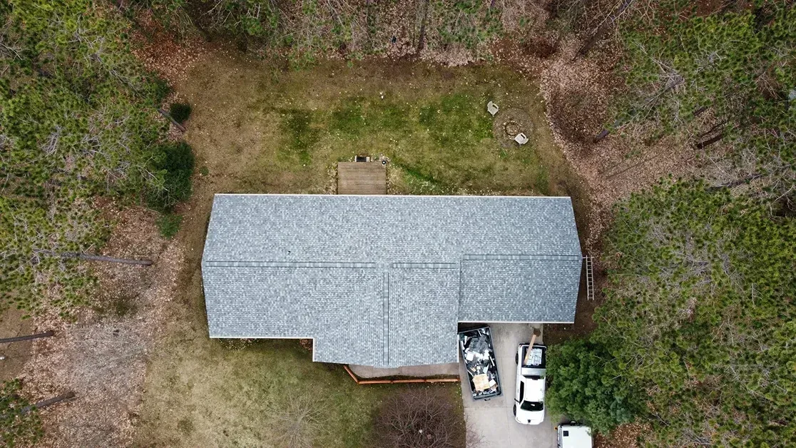 An overhead drone shot of a house with a gray shingled roof, surrounded by trees, with a driveway and parked vehicles.