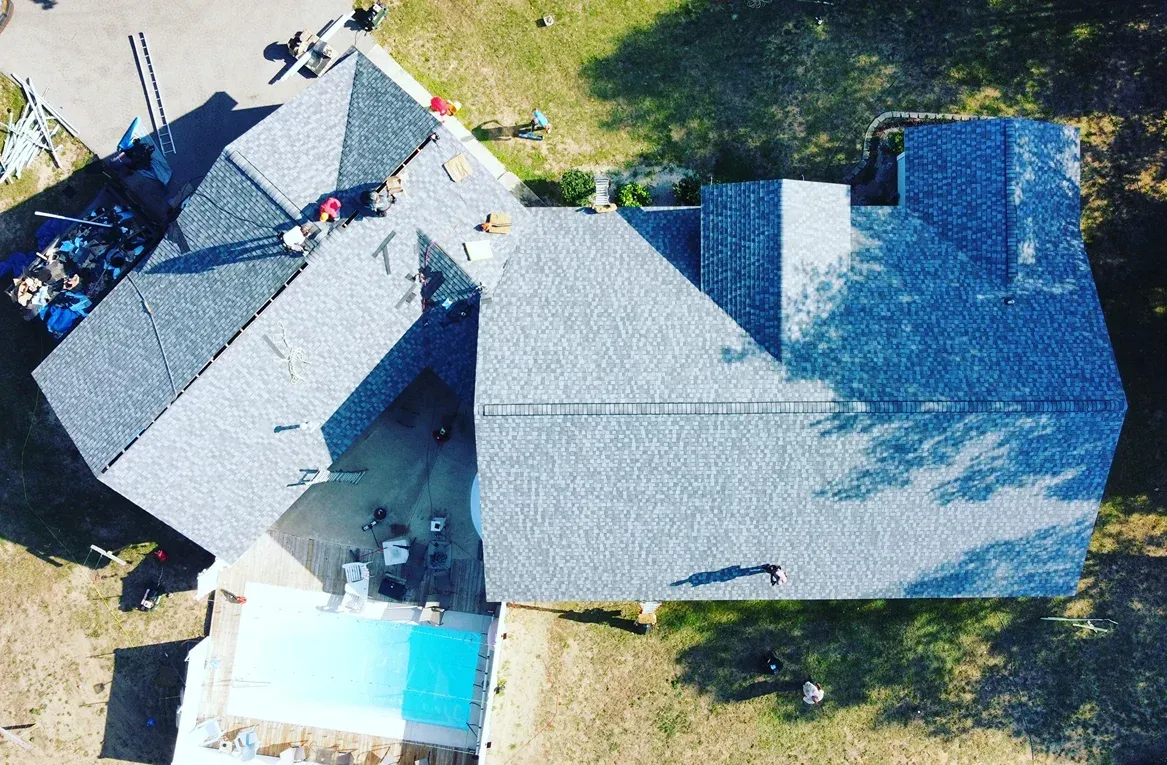 Aerial view of a house with a gray shingled roof under construction, featuring an adjacent swimming pool.