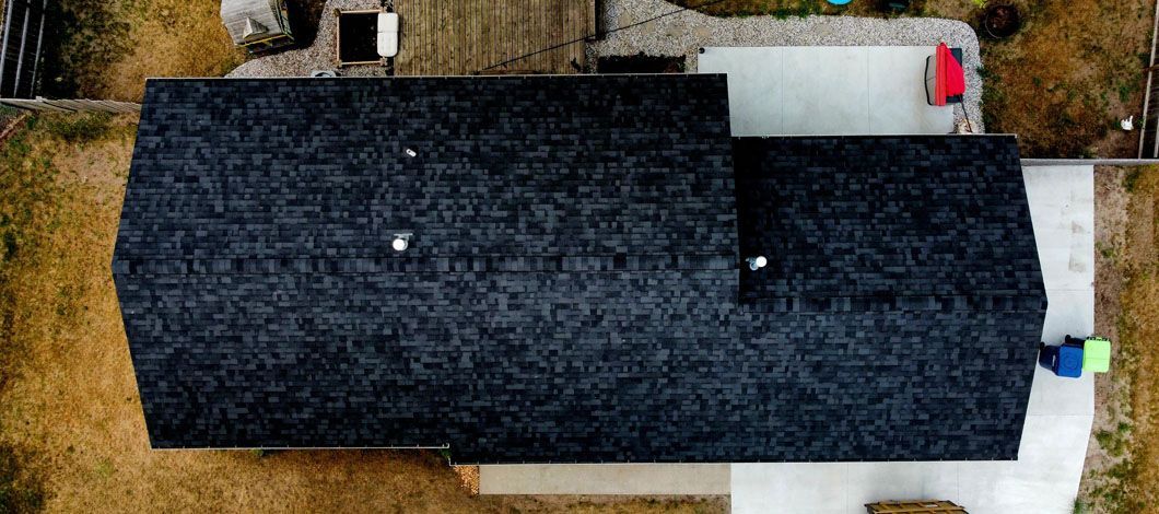 A top-down aerial view of a dark shingled residential roof with two chimneys, surrounded by a light-colored yard.