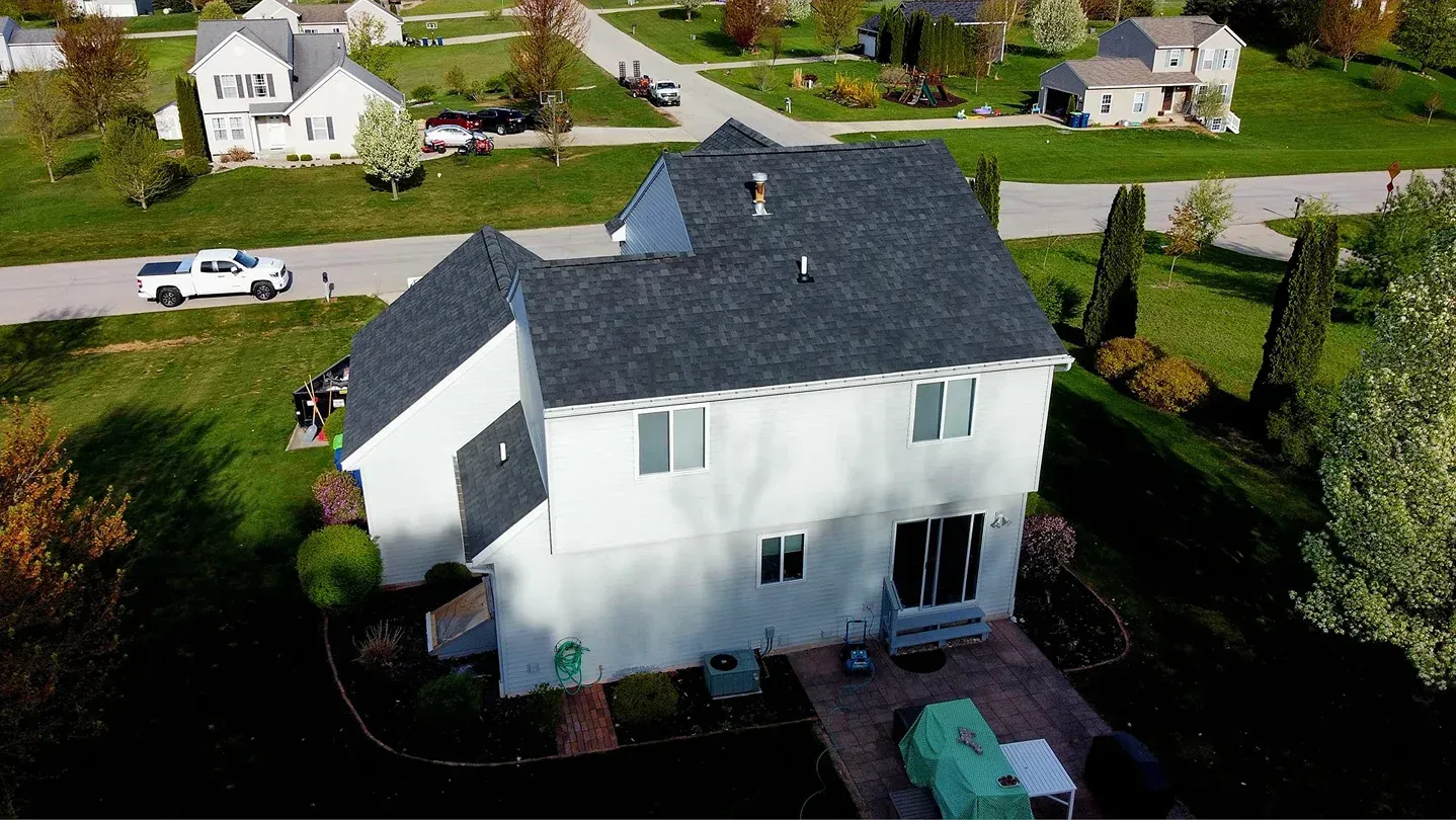 An aerial view of a white two-story house with a dark gray shingled roof in a suburban neighborhood on a sunny day.