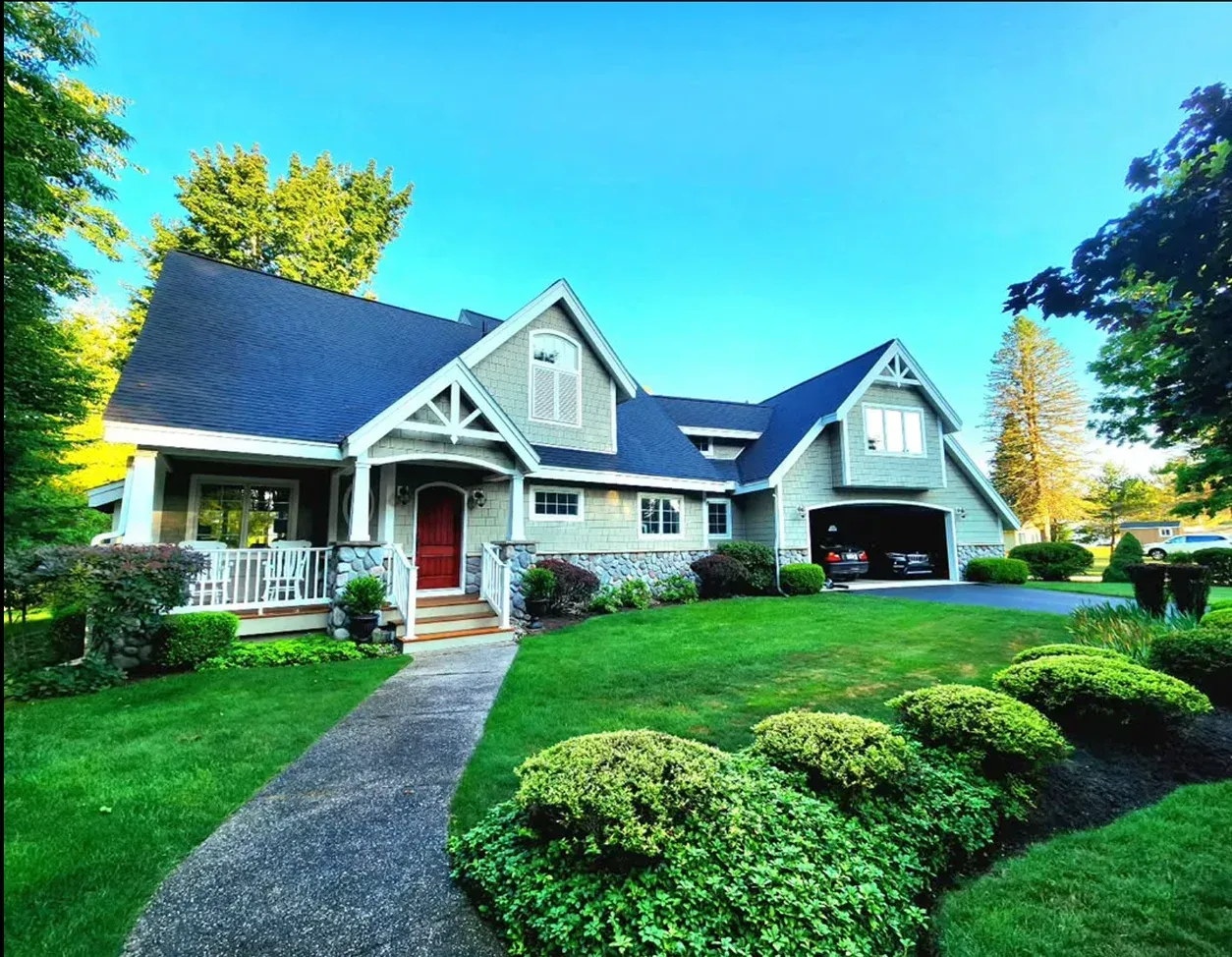 A light gray two-story house with a red front door, blue roof, stone accents, and a landscaped yard under a clear blue sky.