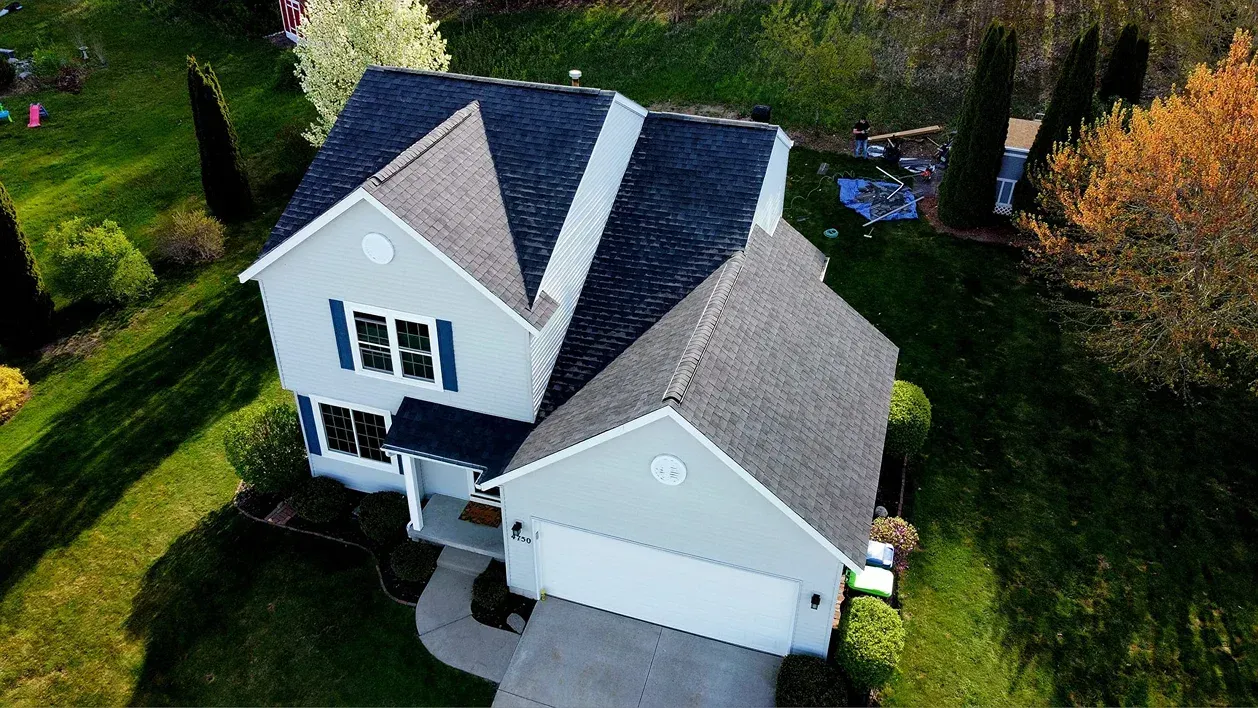 An aerial view of a two-story suburban home with light siding, blue shutters, and a dark shingled roof, surrounded by lawn.