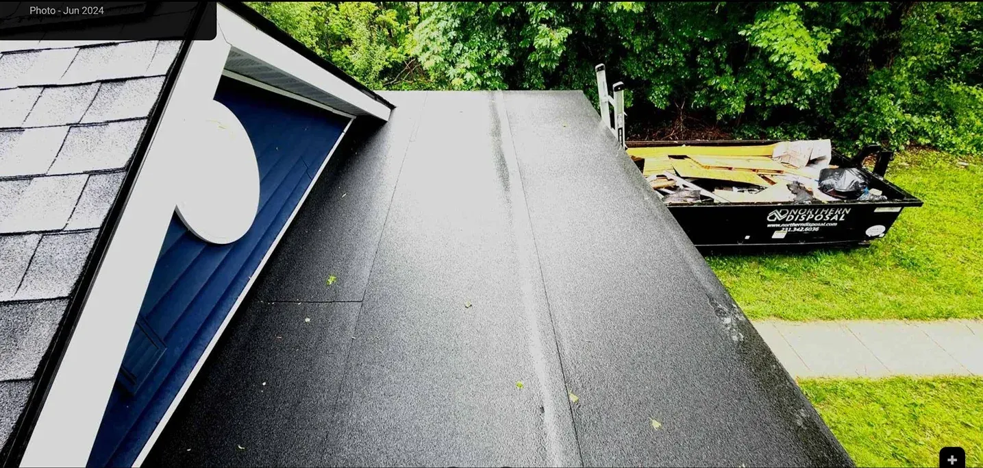 A steep, black rubber roof section meets the side of a blue building with grey shingles on a grassy lot.