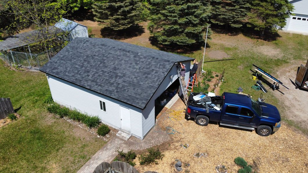 A high-angle view shows a white garage with a new shingled roof, a dark blue truck parked outside, and a person working.