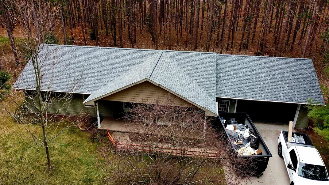 An aerial view of a ranch-style house with a gray shingled roof, a dumpster in the driveway, and a white truck nearby.
