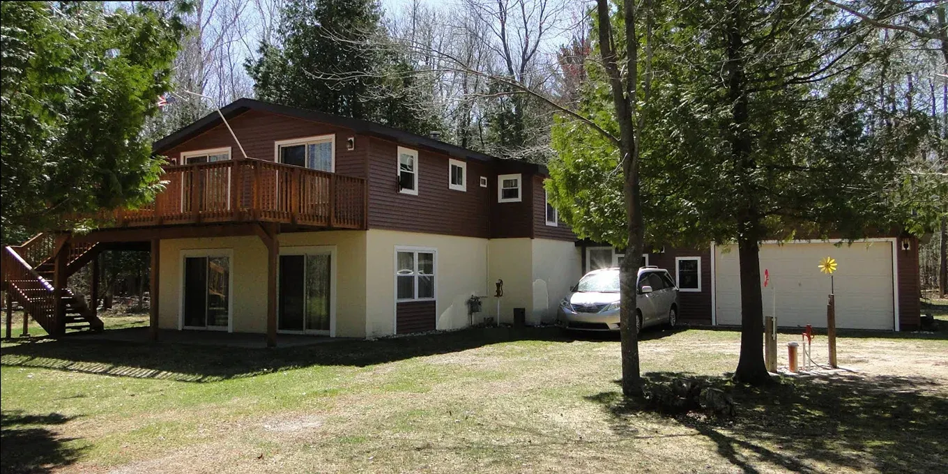 A two-story house with brown wood siding on top and a cream-colored basement level, featuring a wooden deck and garage.