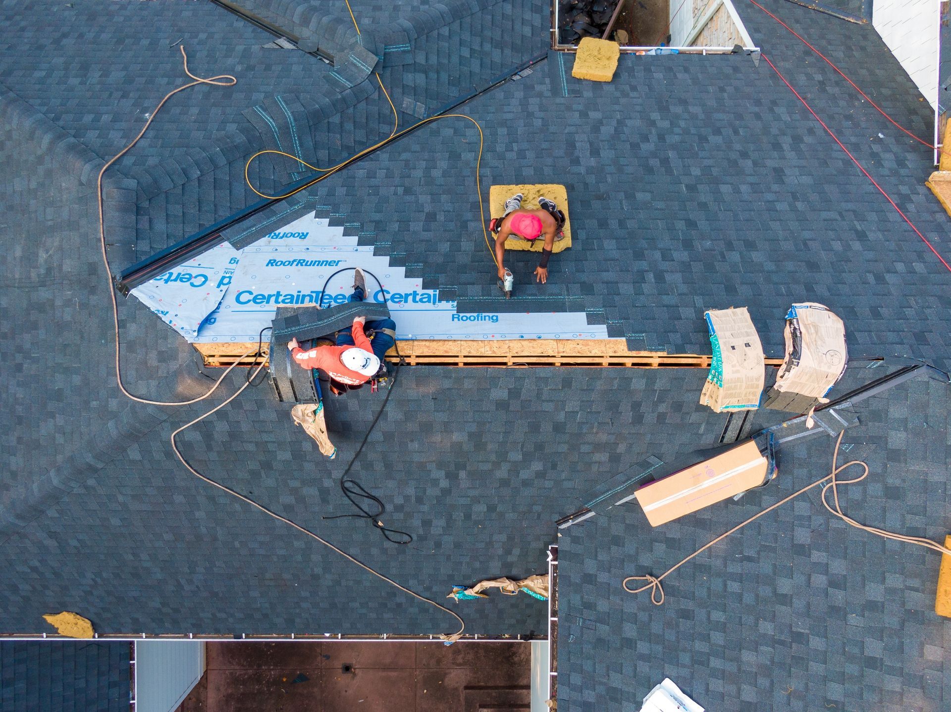 Two workers install dark roof shingles on a house, applying material over light blue CertainTeed underlayment.