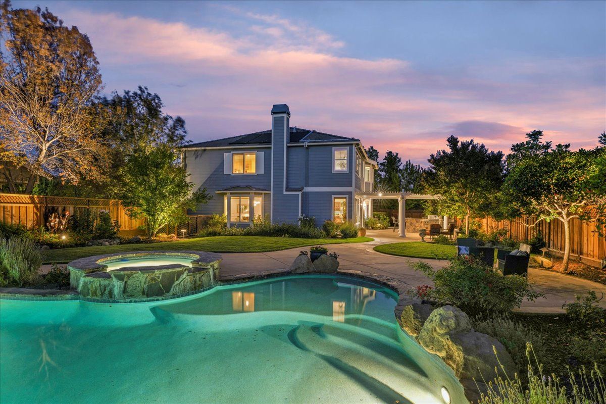 Backyard of a house with a pool, spa, and patio at dusk. Blue exterior, green lawn, trees, and colorful sky.
