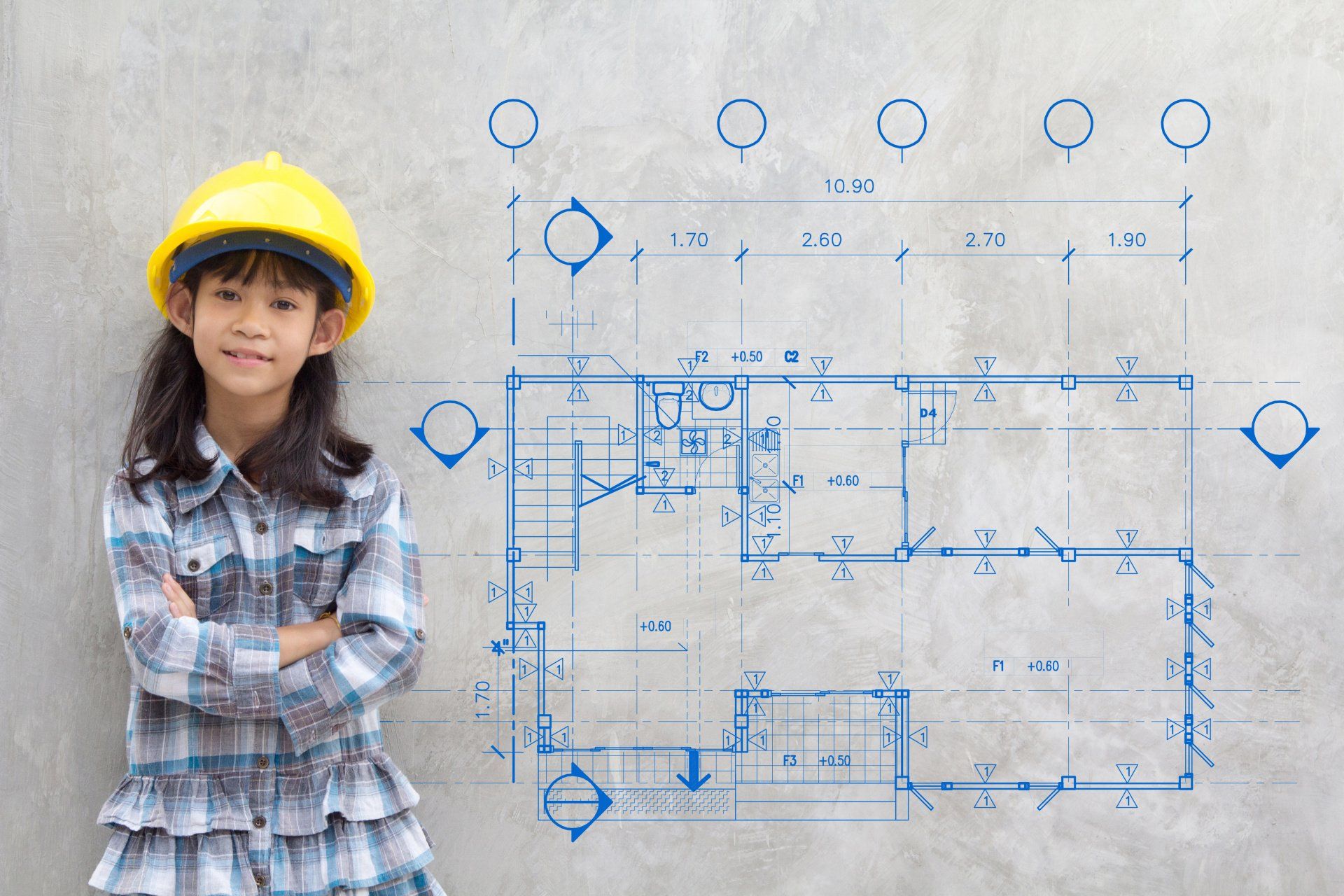 A little girl wearing a hard hat is standing in front of a blueprint.