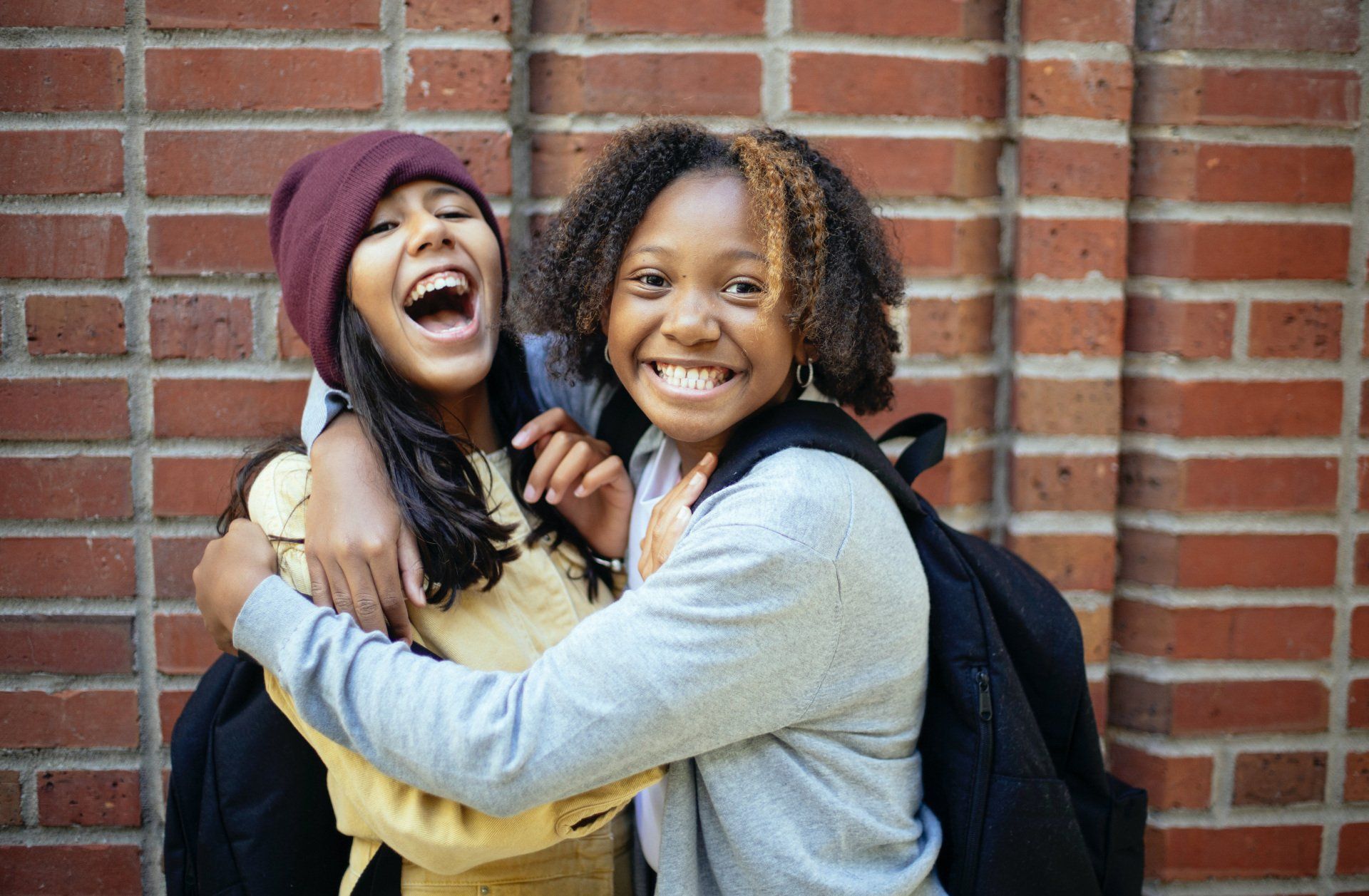 Two girls are hugging each other and laughing in front of a brick wall.
