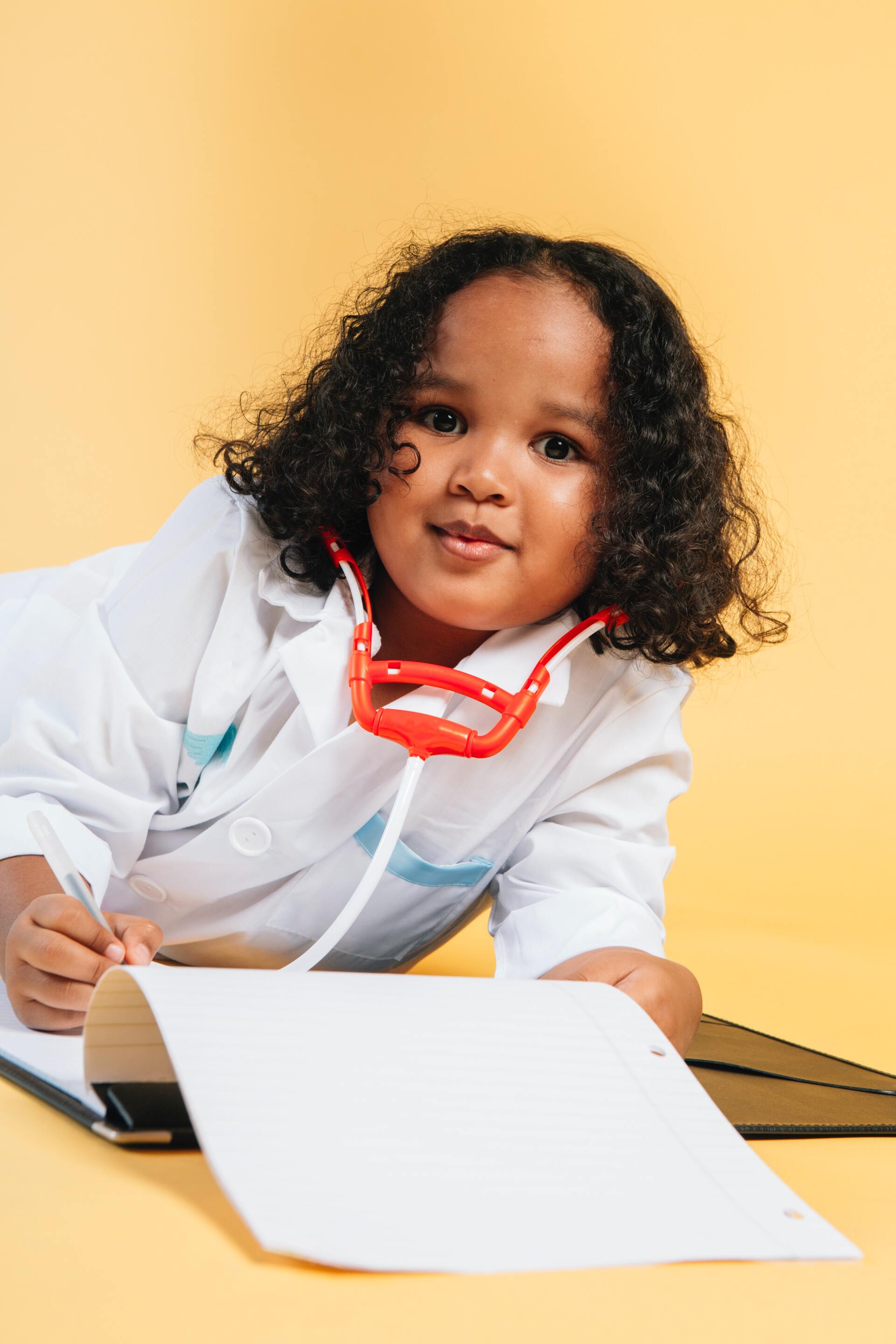 A little girl dressed as a doctor is laying on the floor with a stethoscope around her neck.