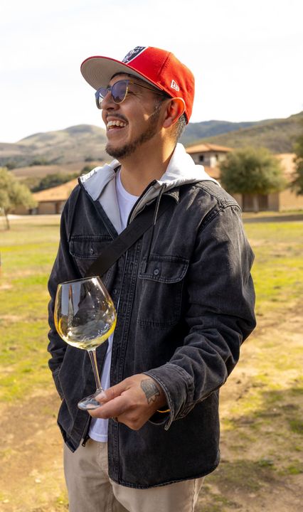 A man wearing a hat and sunglasses is holding a glass of wine.