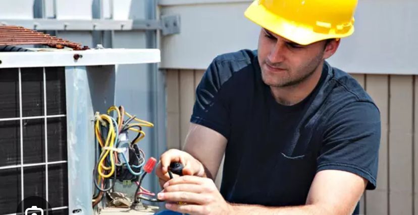 HVAC technician in yellow hard hat repairs an air conditioning unit outdoors, focusing intently.