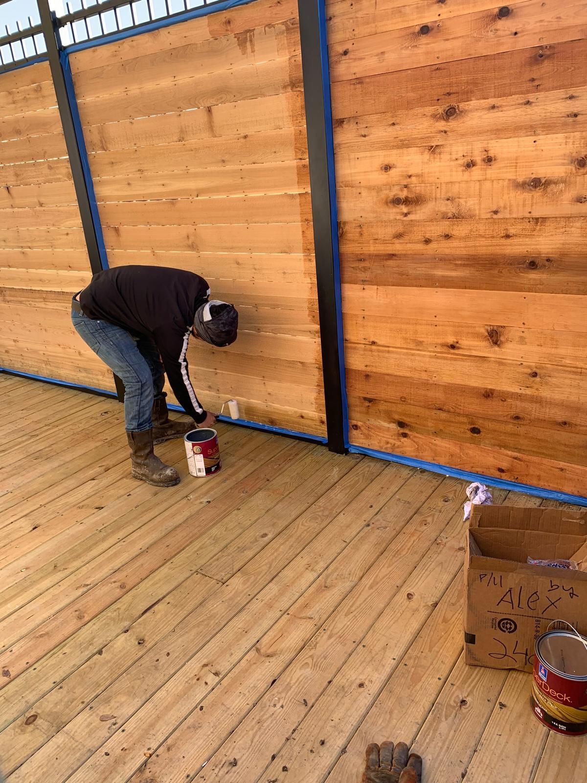 Person painting the base of a wood fence on a wooden deck; blue painter's tape, paint cans visible.