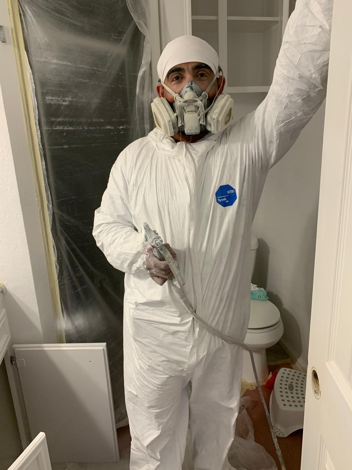 Man in white protective suit and respirator sprays paint in a bathroom.