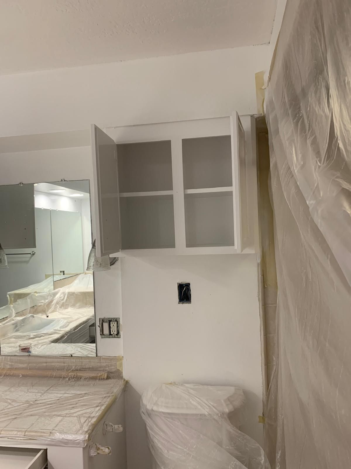 Kitchen cabinet with open door during painting. White wall, cabinet frames, covered countertops.