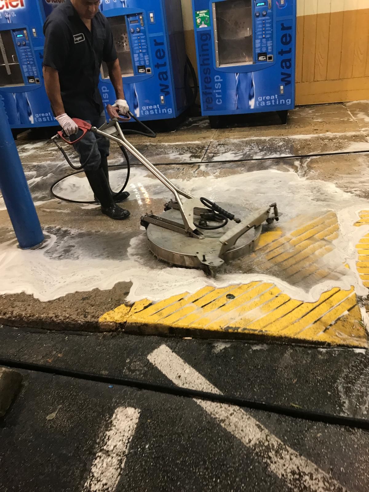 Man using a surface cleaner on a concrete surface, creating a foamy residue near blue water vending machines.