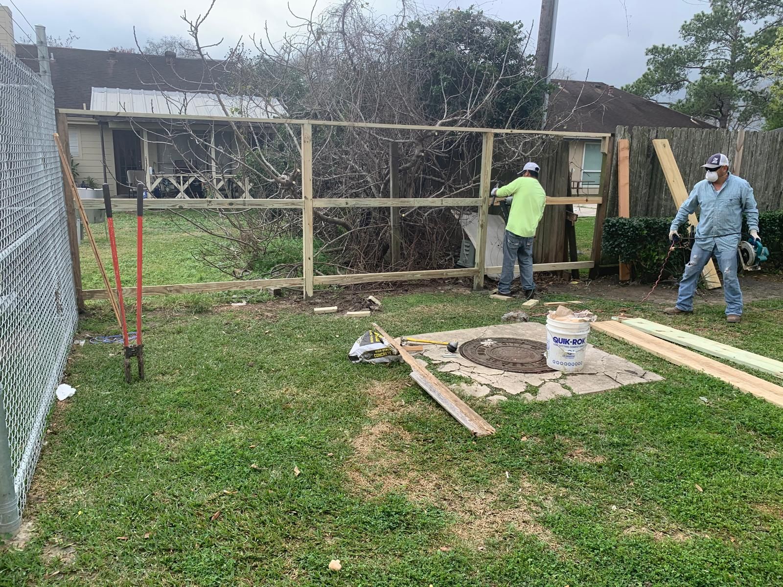 Two men building a wooden fence in a yard. One holds a tool, and the other works on the structure. Green grass, overcast sky.