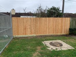 Wooden fence in a backyard with green grass, a chain-link fence, and a concrete utility cover.