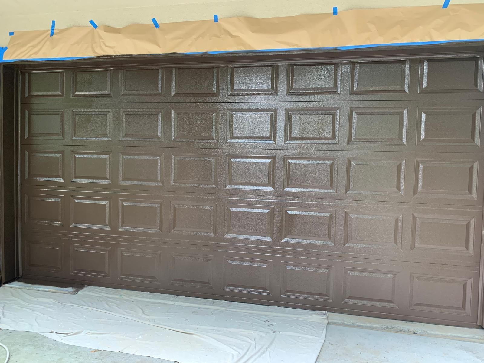 Garage door being painted brown, partially finished. Blue tape and paper protect surrounding areas.