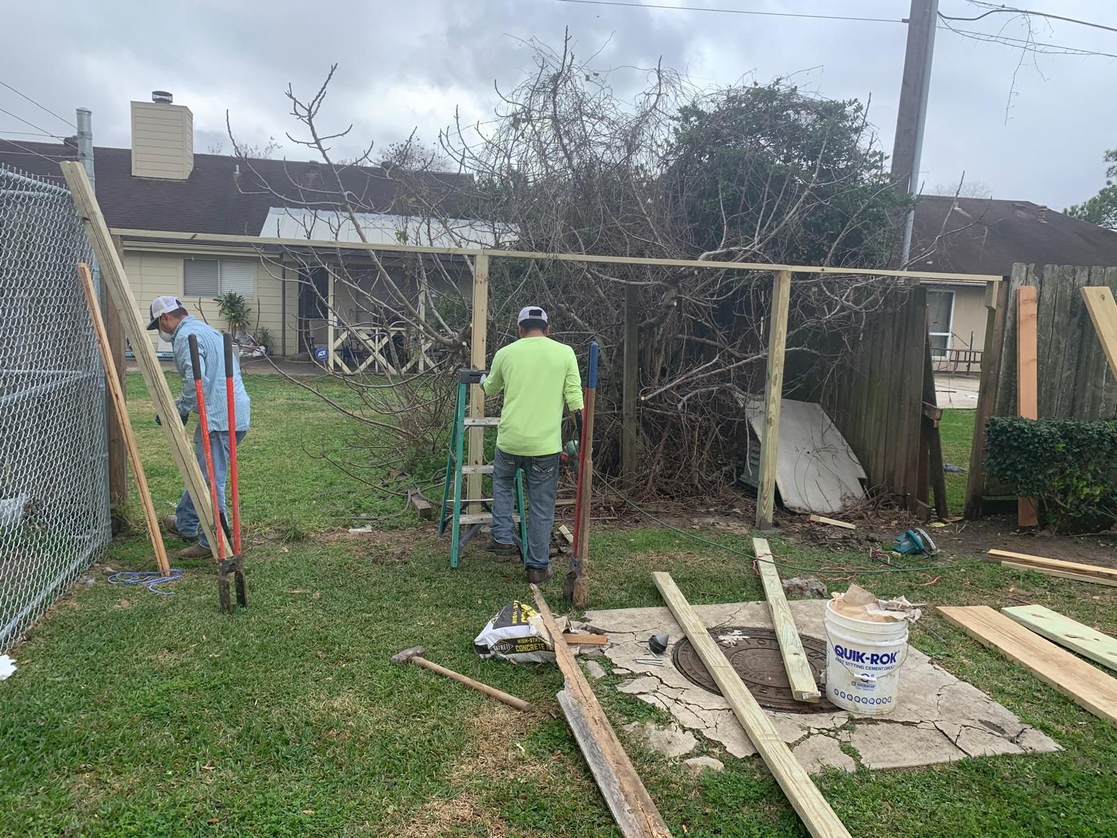 Two men constructing a fence in a backyard with wooden planks and tools, near a chain-link fence and overgrown bushes.