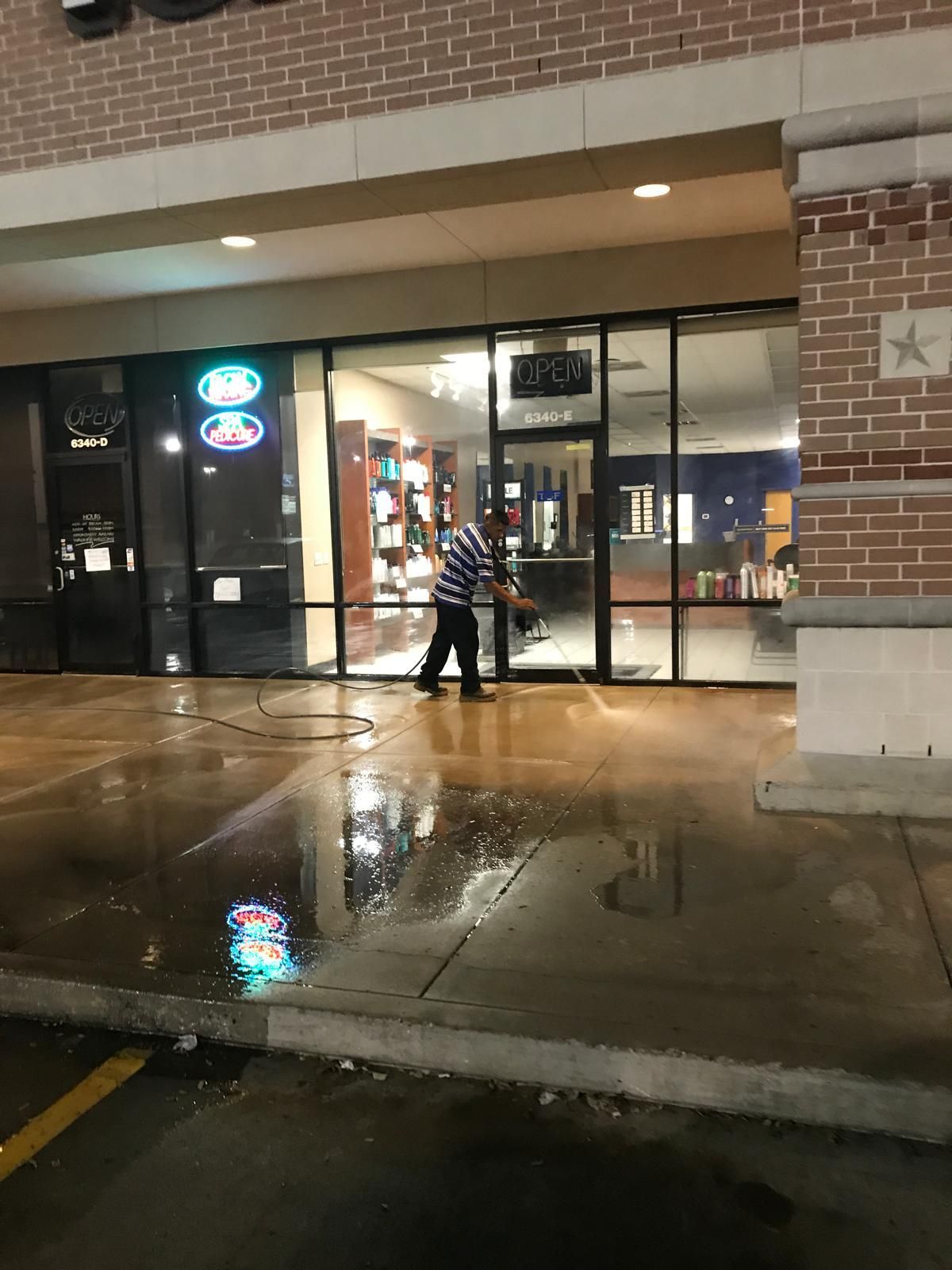 Person sprays water in front of a storefront with large windows. Night. Wet pavement reflects building lights.