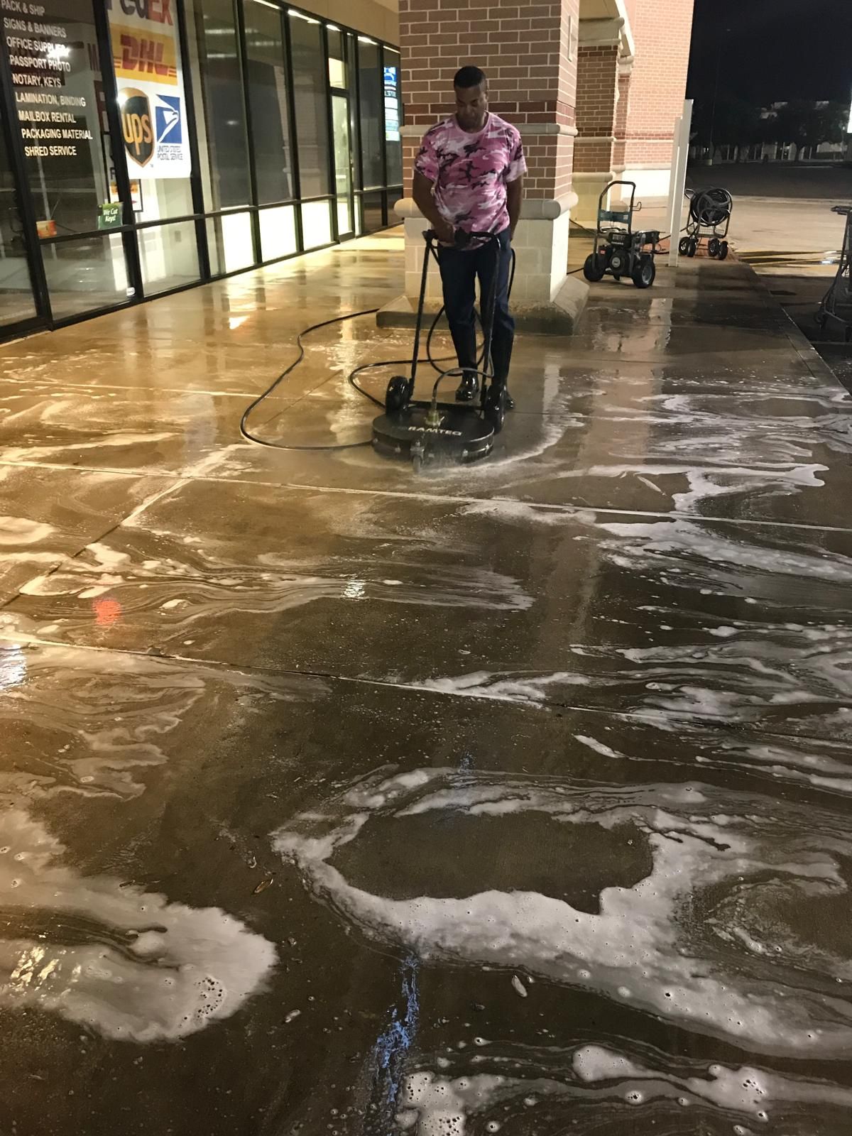 Person pressure washing a commercial sidewalk covered in foamy water at night.