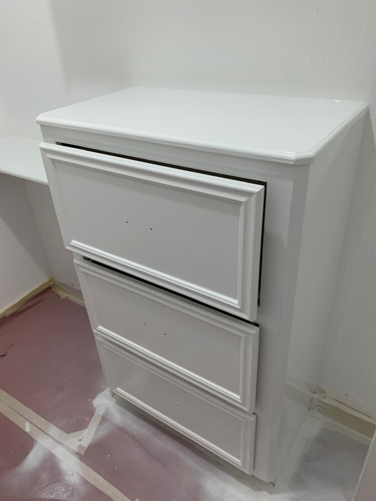 White three-drawer dresser against a white wall. The drawers have decorative panels.