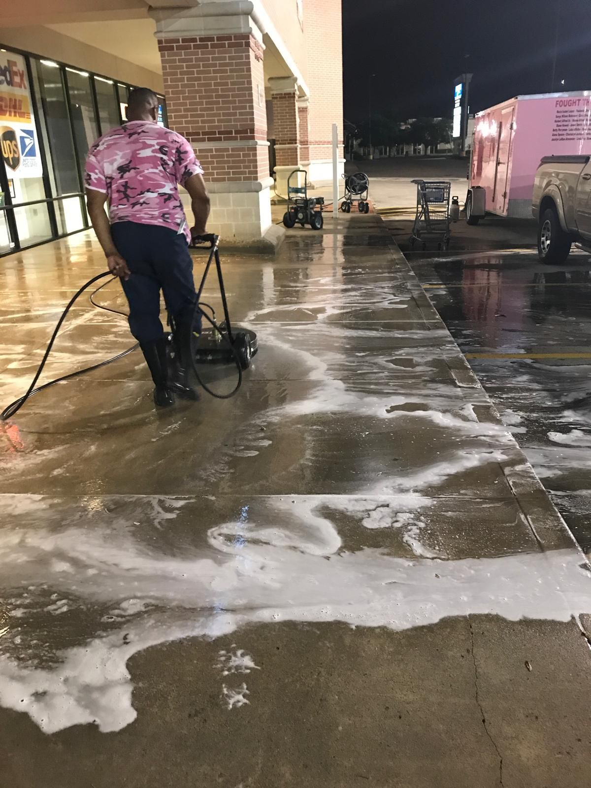 Person in pink camo shirt pressure washing a concrete sidewalk at night.